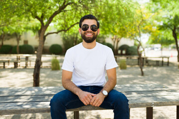 Smiling hispanic man in a white t-shirt and sunglasses sitting on a park bench, ideal for mockups and casual clothing designs - Powered by Adobe