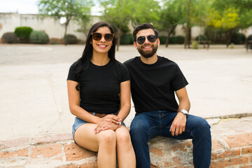 Happy young hispanic couple wearing plain black t-shirts and sunglasses posing for a mockup in a...
