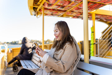 Asian woman using mobile phone making online booking hotel during travel on ferry boat crossing harbor in Sydney, Australia. Attractive girl enjoy outdoor lifestyle travel city on holiday vacation.