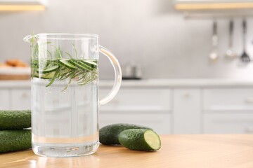 Refreshing cucumber water with rosemary in jug and vegetable on wooden table. Space for text