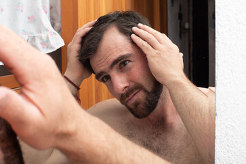worried man looking his hair loss on the mirror, young guy beginning alopecia problem, examining scalp balding distressed
