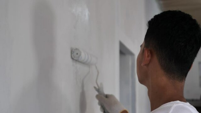 Asian male painter painting new white wall with roller brush, house interior renovation