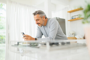 Fototapeta premium Contented mature man with a smartwatch browsing smartphone by a glass-top counter in a sunny kitchen setting