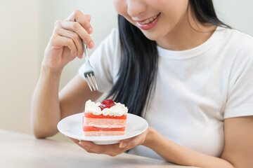 Smiling happy asian young woman holding fork eating slice piece chiffon strawberry layer cake...