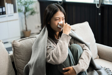 Young Asian woman suffering from flu symptoms, covered with a blanket on a sofa. Concept of illness, healthcare, and recovery