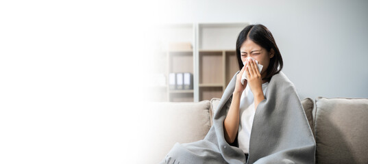 Young Asian woman suffering from flu symptoms, covered with a blanket on a sofa. Concept of illness, healthcare, and recovery