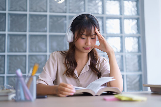 Young asian student in casual clothes wearing headphones to listening the music and reading textbook to thinking ideas while studying education knowledge and doing homework assignments in university