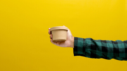 A hand holding a cardboard box against a yellow background, illustrating the concept of shipping,...
