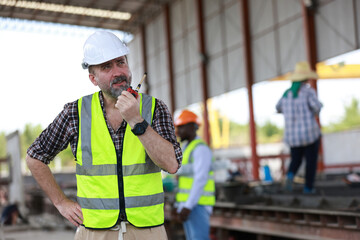 A senior civil engineer is seen from behind while working at a construction site. They are holding a walkie-talkie and overseeing the work progress.