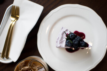 Piece of cheesecake with black currant and blueberry sauce on white plate on wooden table. Cheesecake and cup of coffee on wooden table. Coffee and cake. Horizontal view.