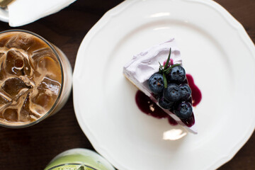 Piece of cheesecake with black currant and blueberry sauce on white plate on wooden table. Cheesecake and cup of coffee on wooden table. Coffee and cake. Horizontal view. top of view.