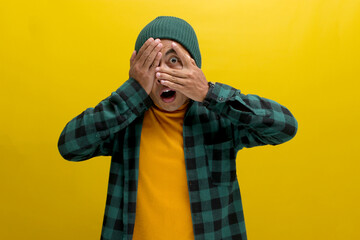 A surprised young Asian man, dressed in a beanie hat and casual shirt, is covering his face and...