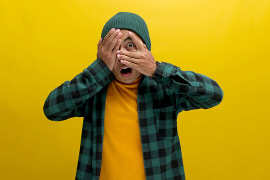 A surprised young Asian man, dressed in a beanie hat and casual shirt, is covering his face and eyes with his hand, peeking through his fingers in a shocked and embarrassed expression