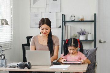 Young mother working on laptop beside her daughter playing on tablet at home office.