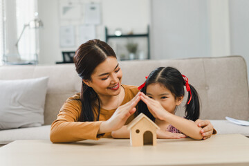 Cute little daughter and mother making symbolic roof of hands above home model together in living room. Safety and protection with love, care and security with happiness at home concept