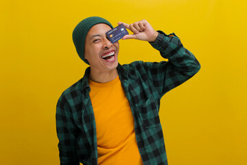An excited young Asian man, wearing a beanie hat and casual shirt, covers one eye with a credit...