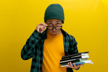 An excited young Asian student, dressed casually in a beanie hat and casual clothes, adjusts his eyeglasses while holding books and looking directly at the camera against a yellow background
