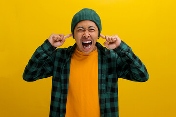 Angry young Asian man, in a beanie hat and casual outfit, is closing his eyes and covering his ears...