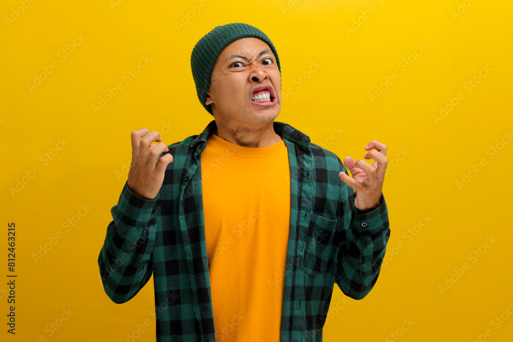 Wall mural Angry young Asian man, dressed in a beanie hat and casual outfit, is shouting and yelling with raised arms, displaying frustration, anger, and agitation while standing against a yellow background