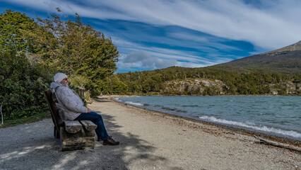 A man in a down jacket rests on an unpainted log bench on the shore of the emerald lake. He's sitting, smiling calmly, looking at the water. The waves are foaming on the pebble beach. Argentina.