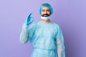 Surgeon man with beard with blue uniform over isolated purple background showing ok sign with fingers