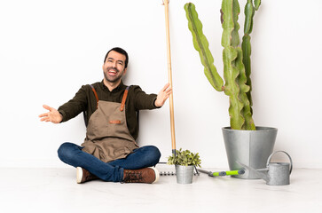 Gardener man sitting on the floor at indoors presenting and inviting to come with hand