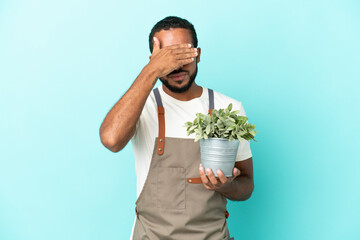 Gardener latin man holding a plant isolated on blue background covering eyes by hands. Do not want...