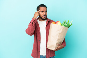 Young latin man holding a grocery shopping bag isolated on blue background having doubts and...