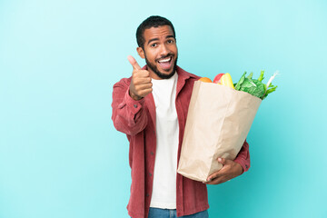 Young latin man holding a grocery shopping bag isolated on blue background with thumbs up because...