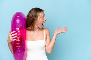 Young blonde woman in swimsuit holding inflatable donut isolated on blue background with surprise...