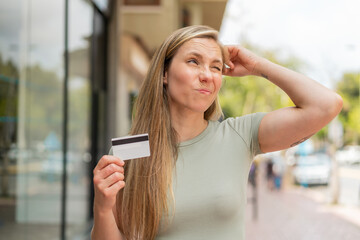 Young blonde woman holding a credit card at outdoors having doubts and with confuse face expression