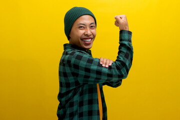 Excited Asian man, dressed in a beanie hat and casual shirt, is making a strong gesture by lifting...