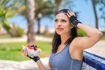 Young pretty sport woman holding a bowl of fruit at outdoors having doubts and with confuse face...