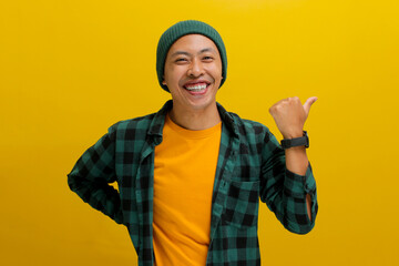 An excited young Asian man, wearing a beanie hat and casual shirt, is smiling while pointing with his thumb to an empty space beside him against a yellow background