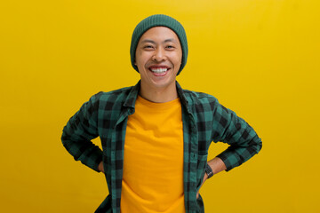 optimistic young Asian man, dressed in a beanie hat and casual shirt, stands confidently with arms...