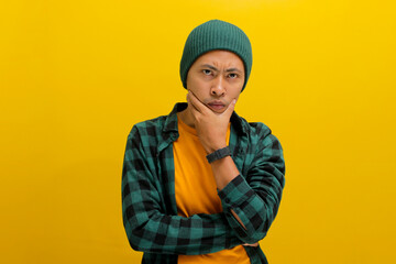 Pensive young Asian man, wearing a beanie hat and casual shirt, places his hand on his chin, looking at the camera with a confused expression, deeply lost in thought and contemplating the future