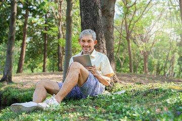 happy asian senior man relaxing under a tree in national park,using digital tablet communicate with family,concept of elderly retired with modern lifestyle,technology,travel,relax