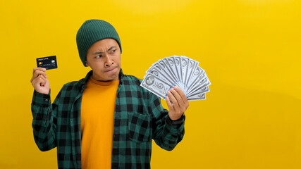 Contemplative Asian man in a beanie and casual clothes examines a credit card and cash, pondering a purchase. Isolated on yellow background. Financial decisions, budgeting, and thoughtful spending