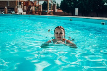 A woman is swimming in a pool. She is smiling and looking at the camera. The pool is blue and the...