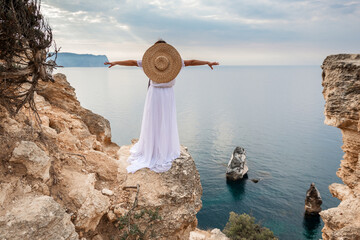 woman white dress stands on a rocky cliff overlooking the ocean. She is wearing a straw hat and she...