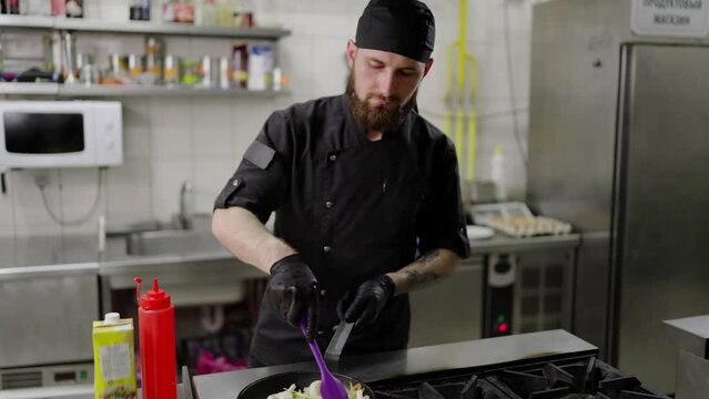 Confident male chef with a beard in a black uniform and with black protective gloves fries vegetables in a frying pan adding oil in a modern kitchen in a restaurant