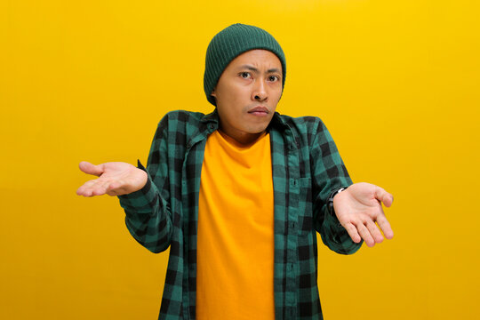 Young Asian man, dressed in a beanie hat and casual shirt, is making an I don't know Italian gesture, with open arms, raised shoulders, and showing both palms while standing against yellow background.