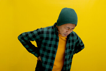 Young Asian man, dressed in a beanie hat and casual outfit, is shown holding his lower back, visibly experiencing unbearable pain due to a backache, against a yellow background.