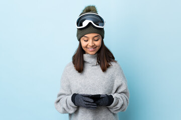 Mixed race skier girl with snowboarding glasses over isolated blue background sending a message...