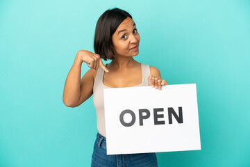 Young mixed race woman isolated on blue background holding a placard with text OPEN and  pointing it