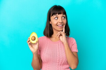 Young mixed race woman holding an avocado isolated on blue background thinking an idea while...