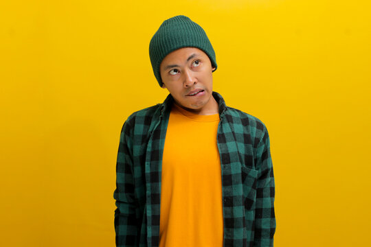 Hesitant young Asian man, dressed in a beanie hat and casual shirt, bites his lips, conveying a playful oops moment as he looks away towards empty space, against a yellow background