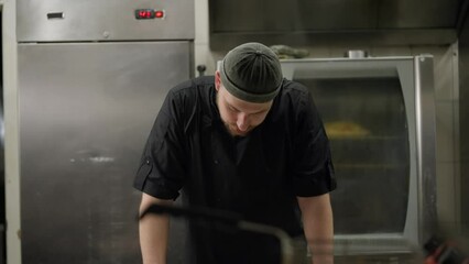 A tired sad cook in a black uniform takes off his hat during a hard day at work and leans his elbows on the table trying to rest during a hard day at work in the kitchen of a restaurant