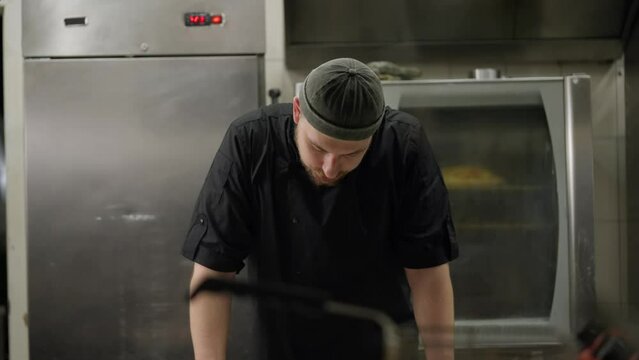 A tired sad cook in a black uniform takes off his hat during a hard day at work and leans his elbows on the table trying to rest during a hard day at work in the kitchen of a restaurant