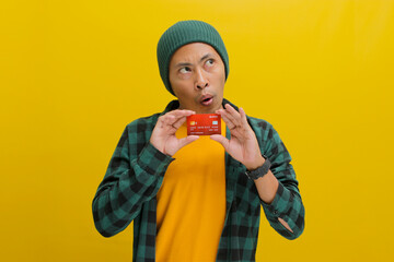 A surprised young Asian man, dressed in a beanie hat and casual shirt, holds a credit card while standing against a yellow background